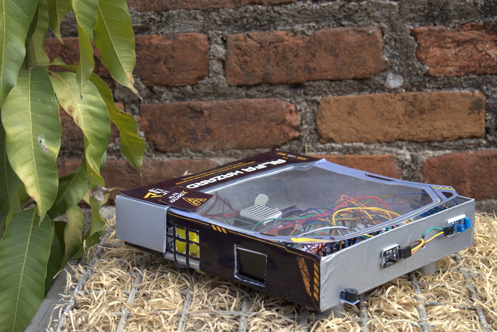 The Alfa HazardX v1 reference build outdoors on a brick ledge: a hand-built enclosure with a clear lid over the board, a dot-matrix display and a coloured wiring loom, with the gas-sensing elements mounted through the side wall.